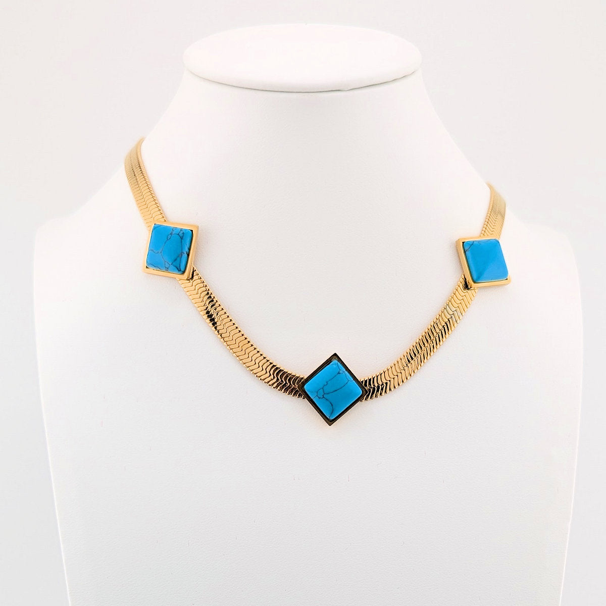 A gold-tone chain necklace displayed on a white mannequin bust, featuring three square-shaped bright turquoise blue stone inlays set in gold at the chain's points.