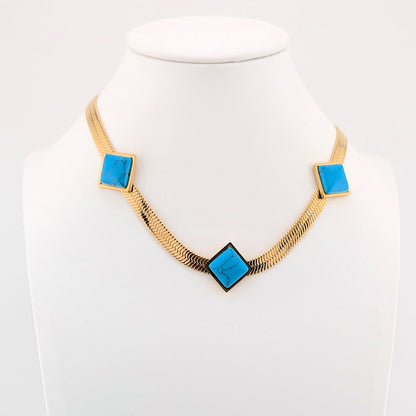 A gold-tone chain necklace displayed on a white mannequin bust, featuring three square-shaped bright turquoise blue stone inlays set in gold at the chain's points.