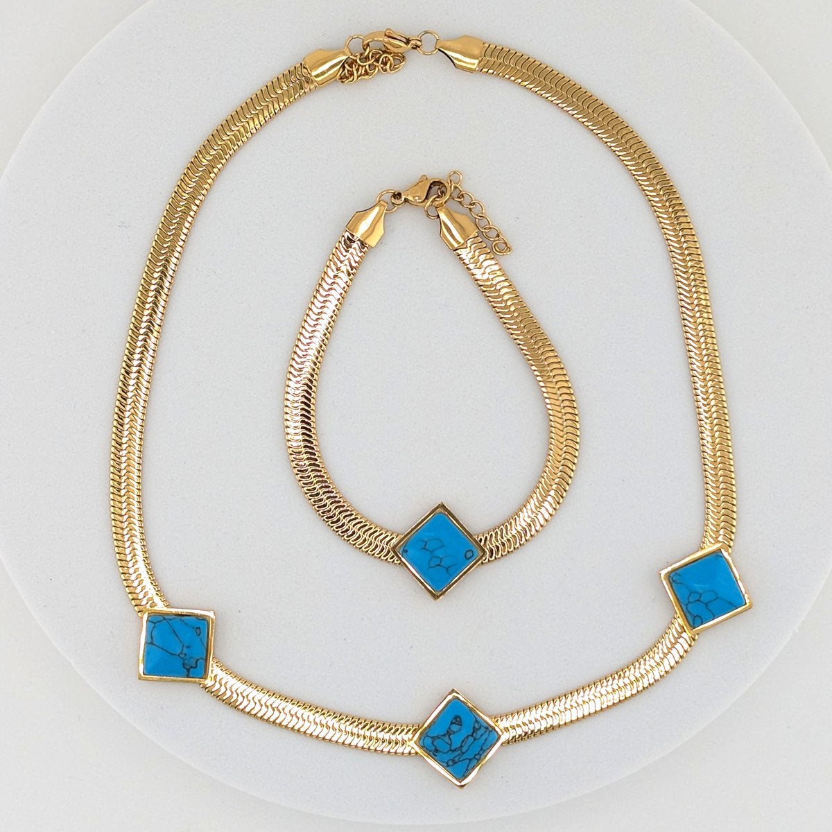 Gold snake chain necklace and matching bracelet set, each decorated with three square turquoise blue inlay accents, displayed on a white circular surface.