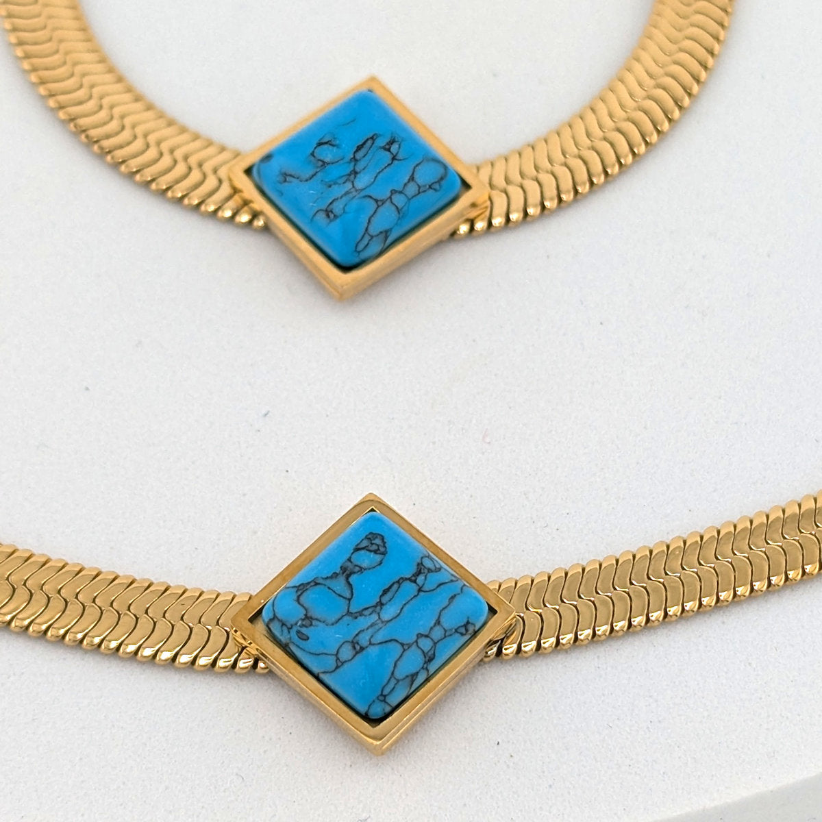 Gold snake-link chain necklace with two square turquoise inlays featuring black marbling, displayed on a white surface.