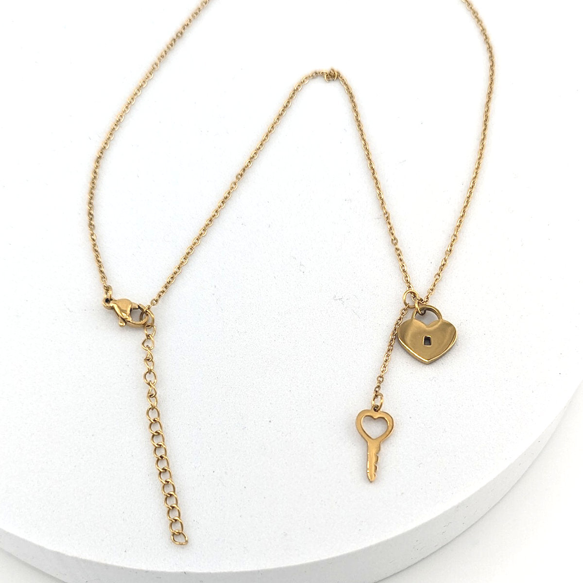 A delicate gold necklace with a heart-shaped padlock and key pendant, displayed on a white circular pedestal.